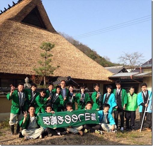 法被姿の篠山東雲高等学校の生徒らがのぼり旗とVサインをして茅葺屋根の建物の前で記念撮影している写真